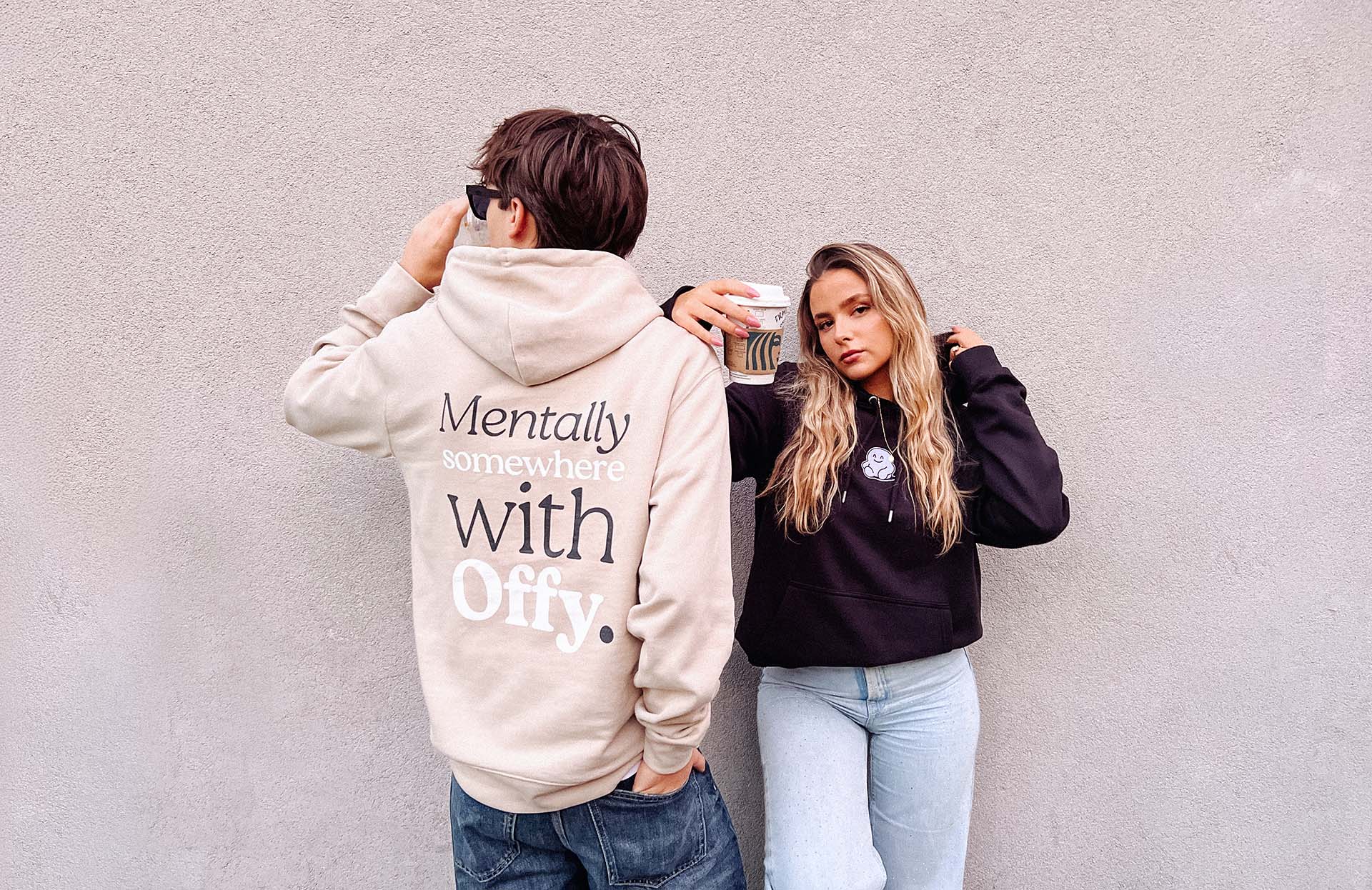Two people wearing hoodies with text, standing against a plain wall.